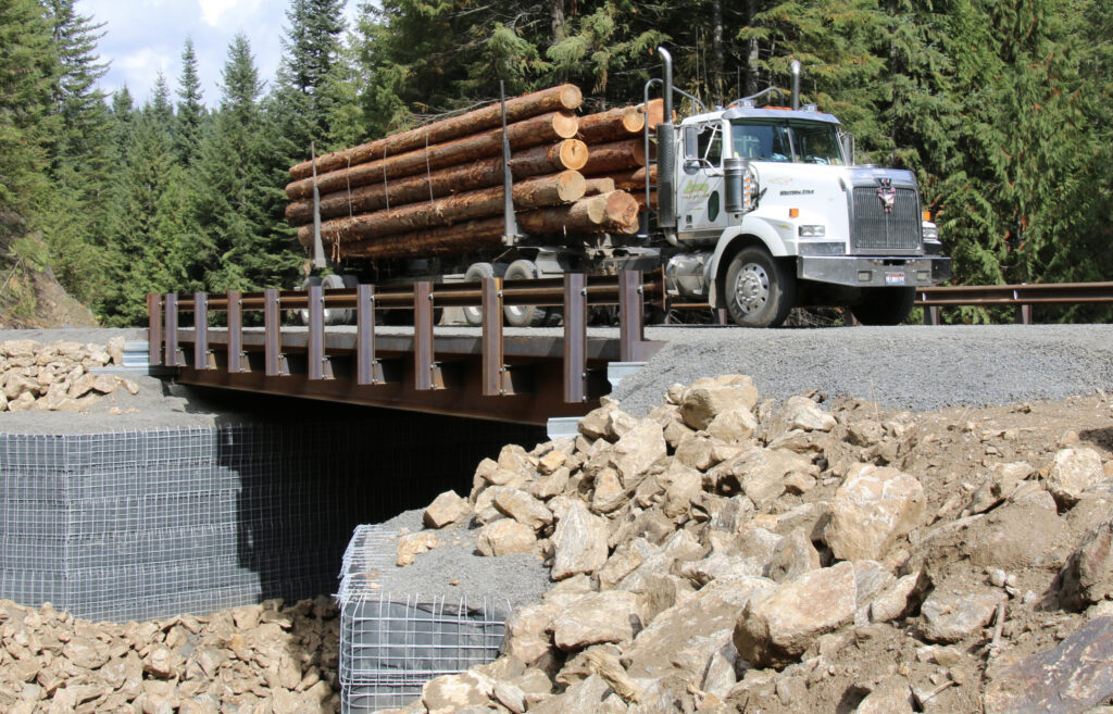 tractor trailer loaded with logs crossing a stream on a steel grider bridge